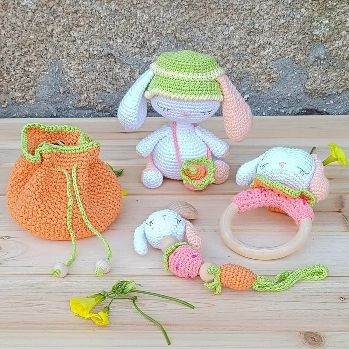 Set en crochet para bebé PomPóm con peluche de apego, sonajero de madera, chupetero y guirnalda sobre fondo neutro.